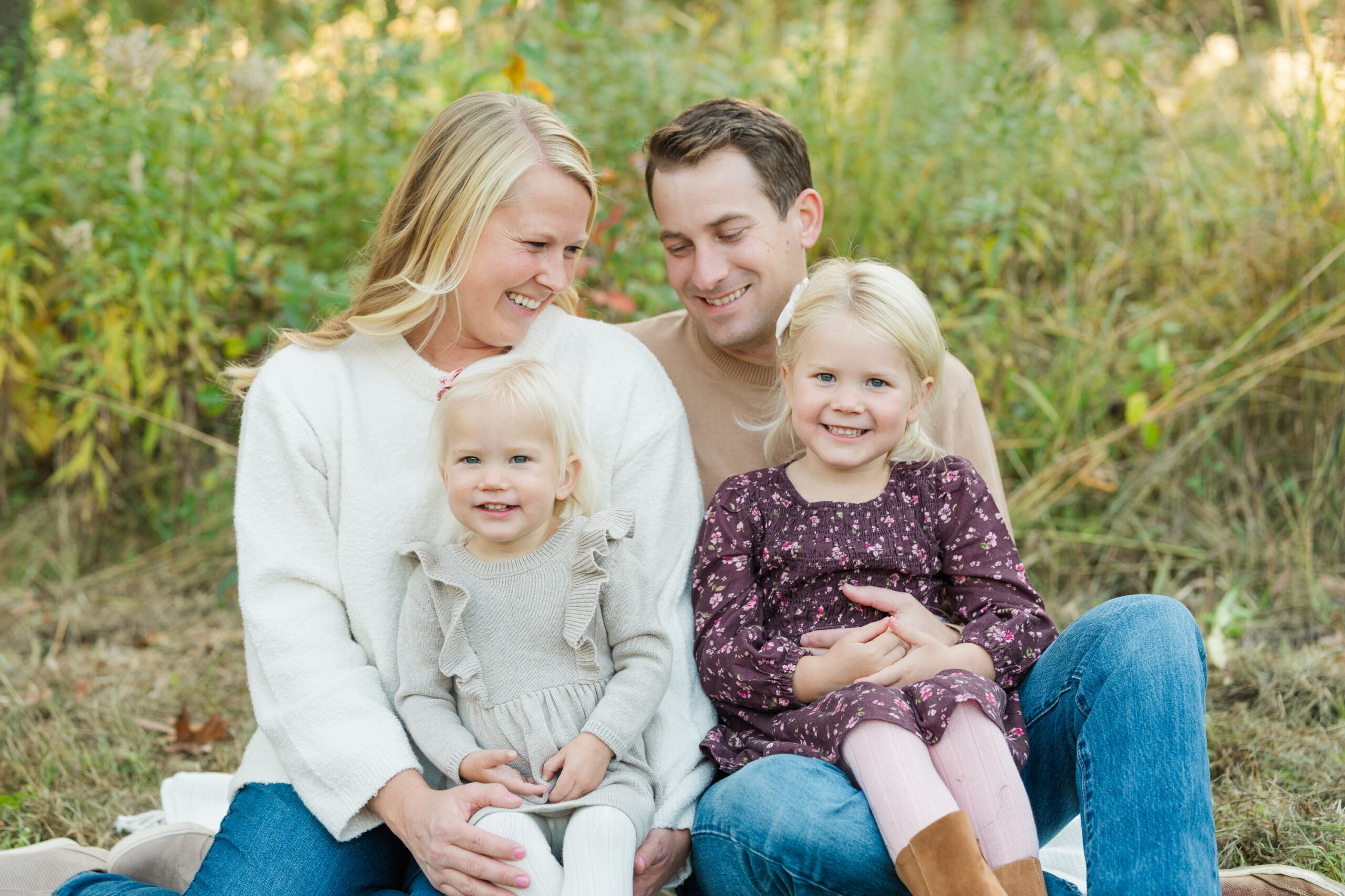 Family of four sitting close together on a blanket in a grassy field, smiling and enjoying a quiet moment surrounded by fall foliage.