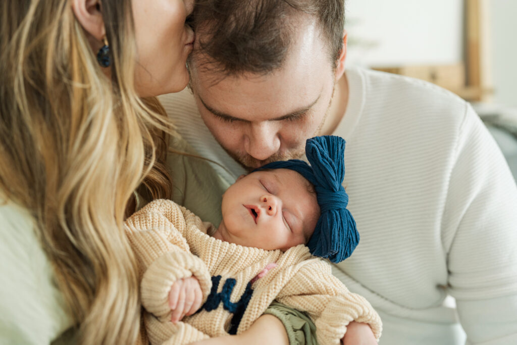 Parents kissing newborn baby girl wearing blue bow during in home newborn photography session in Minnesota.