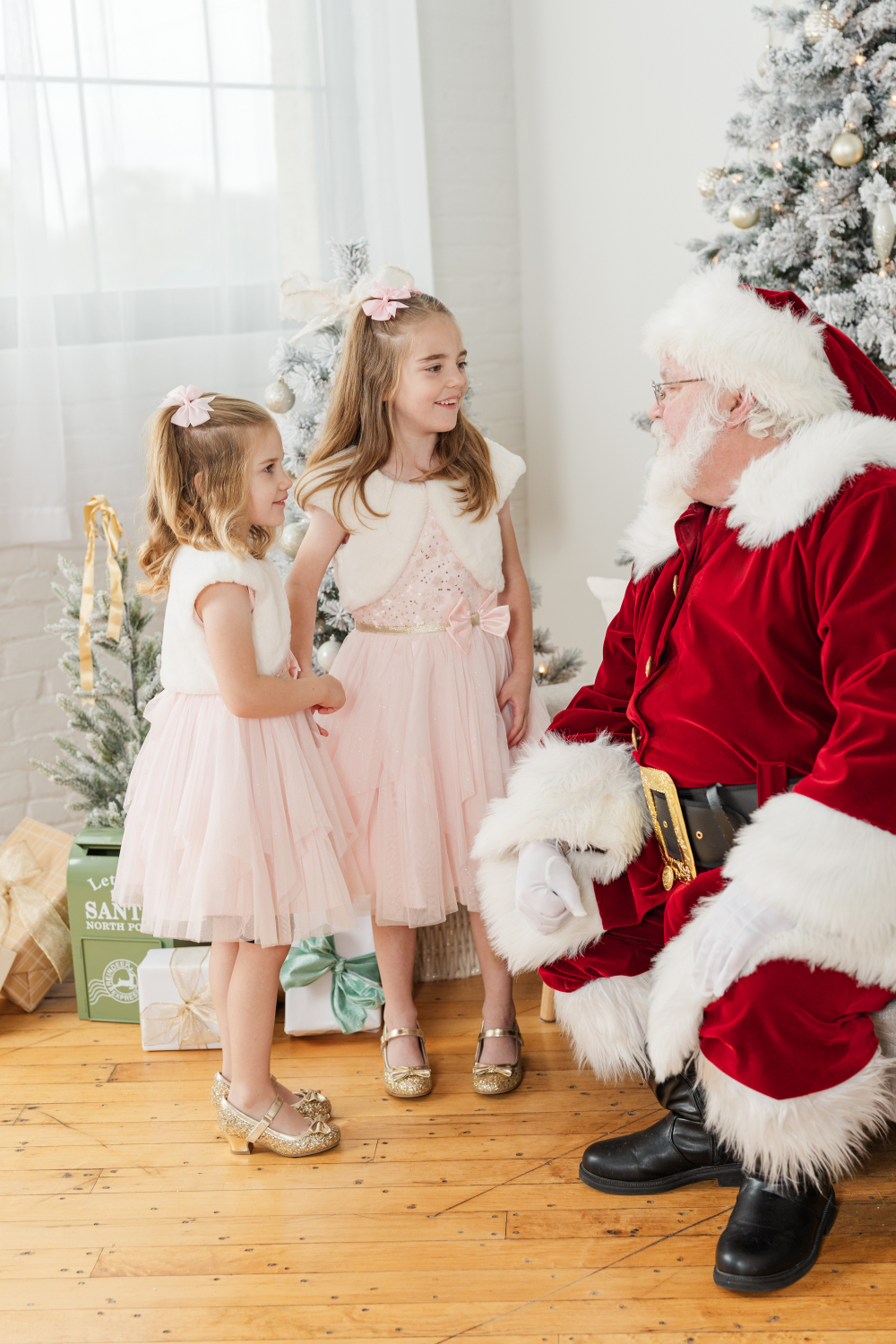 Two smiling girls in matching holiday dresses chatting with Santa in a cozy, beautifully decorated Christmas studio.