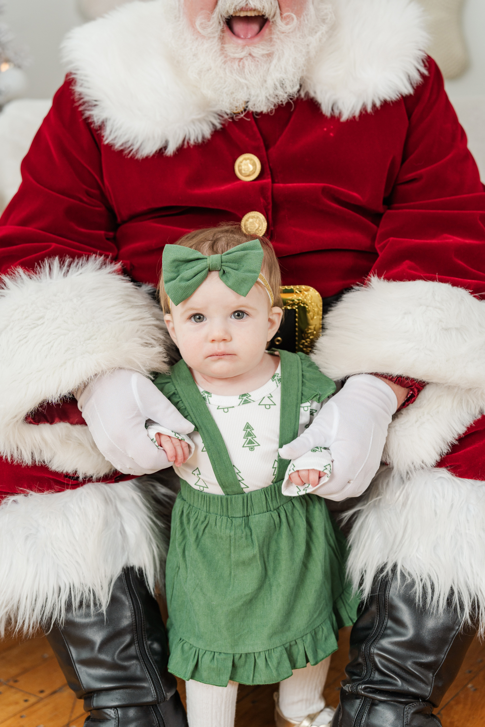 Baby in green holiday outfit sitting on Santa’s lap, looking sweet and curious in a festive Christmas setting.