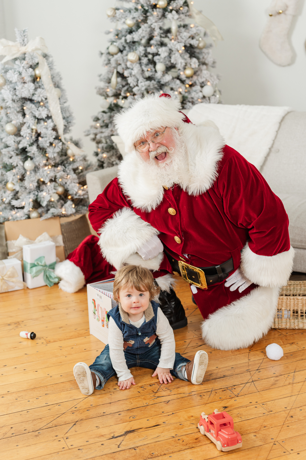 Santa kneeling beside toddler during professional Santa photos in Minneapolis holiday studio with trees and toys.