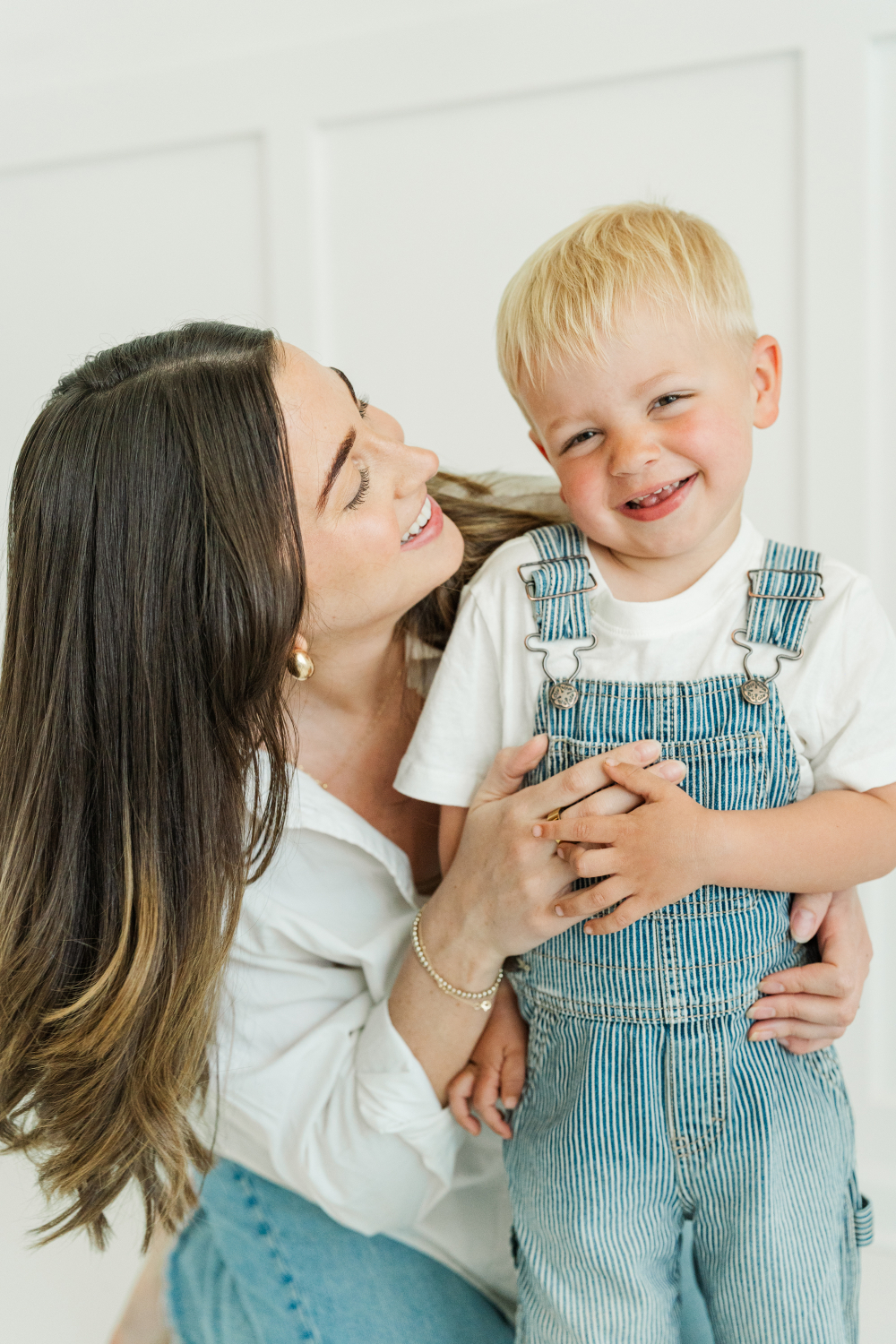 Mother smiling up at her toddler son in striped overalls, sharing a playful and tender moment.