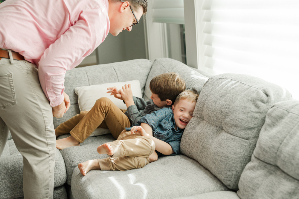 Two young brothers laughing and cuddling on a couch during a playful, light-filled family moment.
