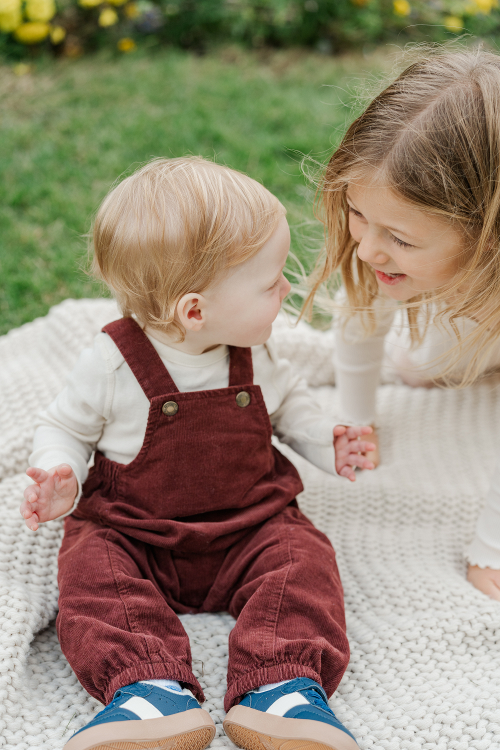 Brother and sister share a joyful moment on a blanket outside, smiling and holding hands on a cozy day.