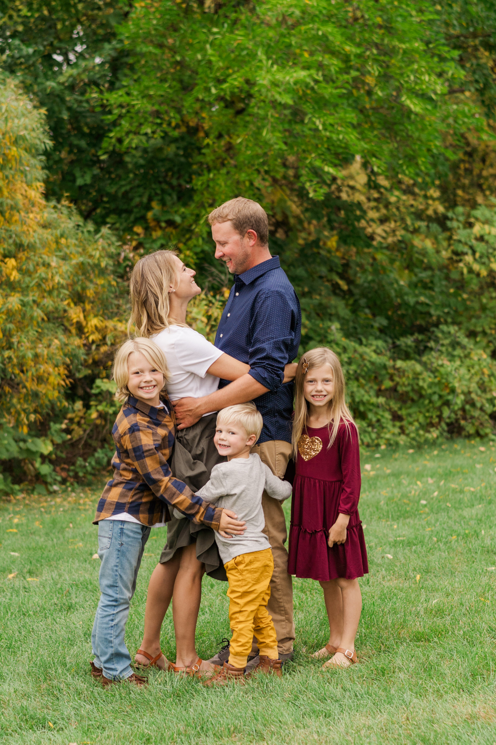 Family of five hugging and smiling together outdoors with fall trees in background.