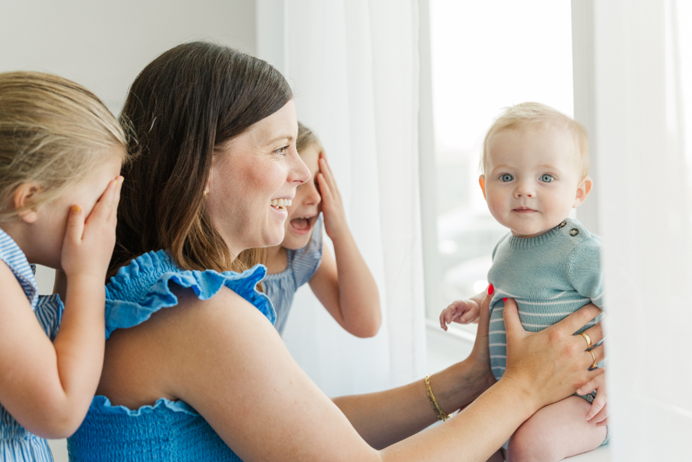Mother smiling at baby by window while daughters laugh beside her indoors.