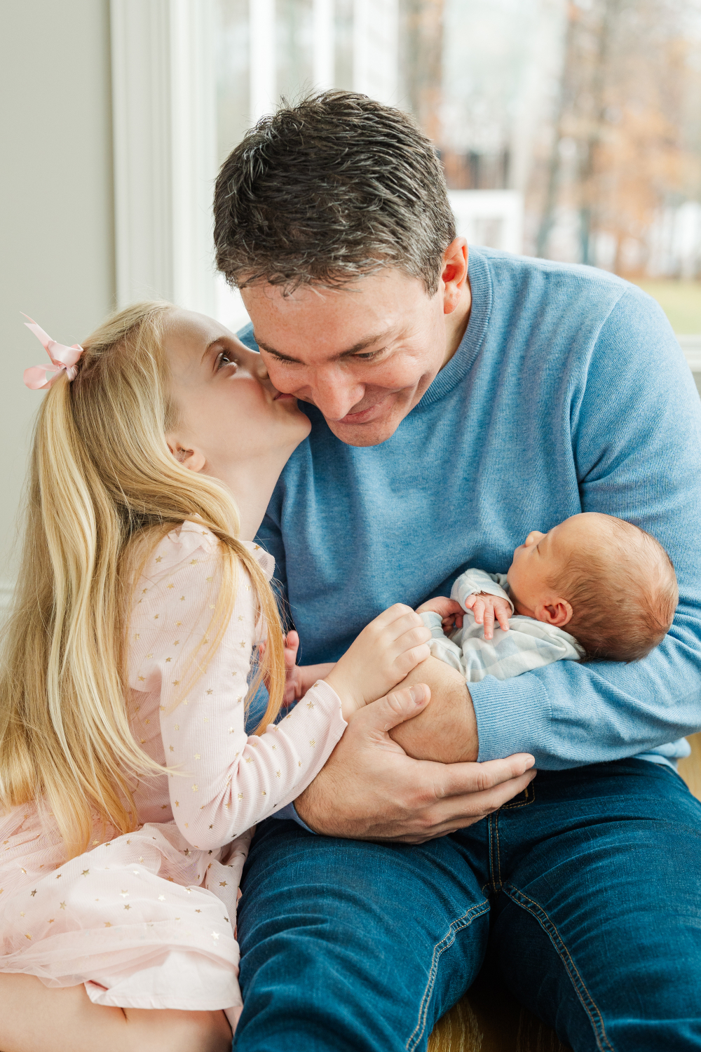 Father holds sleeping newborn while daughter leans in to give him a kiss.