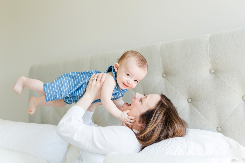 Mother lying on bed lifting smiling baby into the air during a playful, light-filled moment.