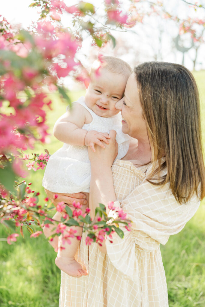 Mother holding happy baby surrounded by pink blossoms in a soft, light-filled spring portrait.