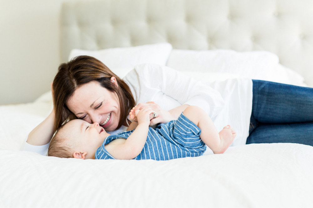 Mother cuddling and smiling at baby while lying together on a soft white bed.