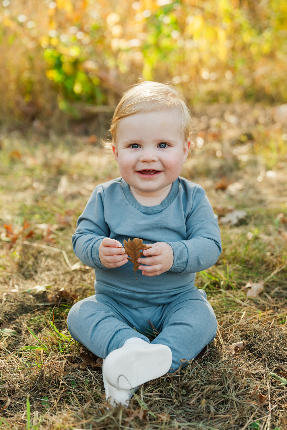 Smiling baby holding a leaf while sitting in soft fall grass, warm and natural outdoor moment