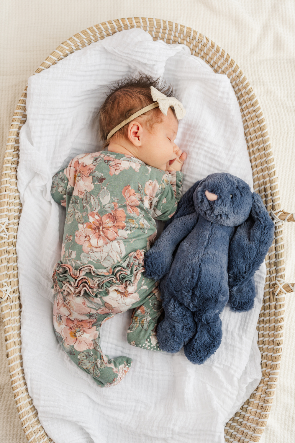 Baby girl sleeps peacefully in a woven bassinet beside a soft blue bunny while wearing a floral sleeper and bow headband. This cozy image beautifully showcases timeless newborn photoshoot outfits with soft textures, muted florals, and natural styling perfect for newborn photography.