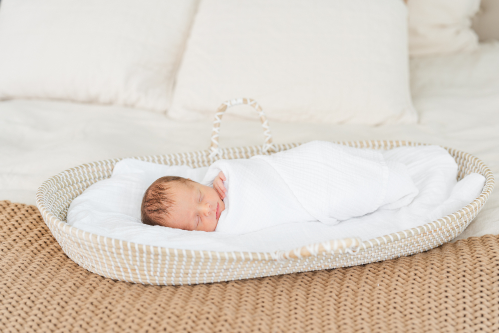 Tiny newborn baby sleeps peacefully in a woven Moses basket wrapped in a soft white swaddle during a bright in-home session. This natural setup beautifully showcases simple newborn photoshoot outfits and neutral styling that create soft, timeless newborn photographs.
