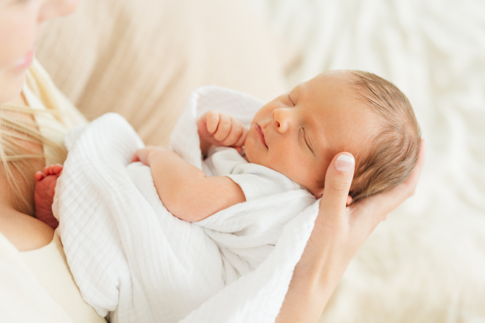 Mother gently cradles her sleeping newborn wrapped in a soft white swaddle during an emotional in-home session. This heartfelt image highlights how simple newborn photoshoot outfits and natural textures help create timeless and connected family photographs.
