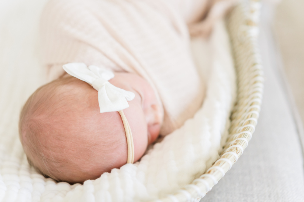 Newborn baby girl rests peacefully in a woven basket while wearing a delicate white bow headband and soft neutral wrap. This light and airy image showcases classic newborn photoshoot outfits that feel timeless, natural, and beautifully simple.