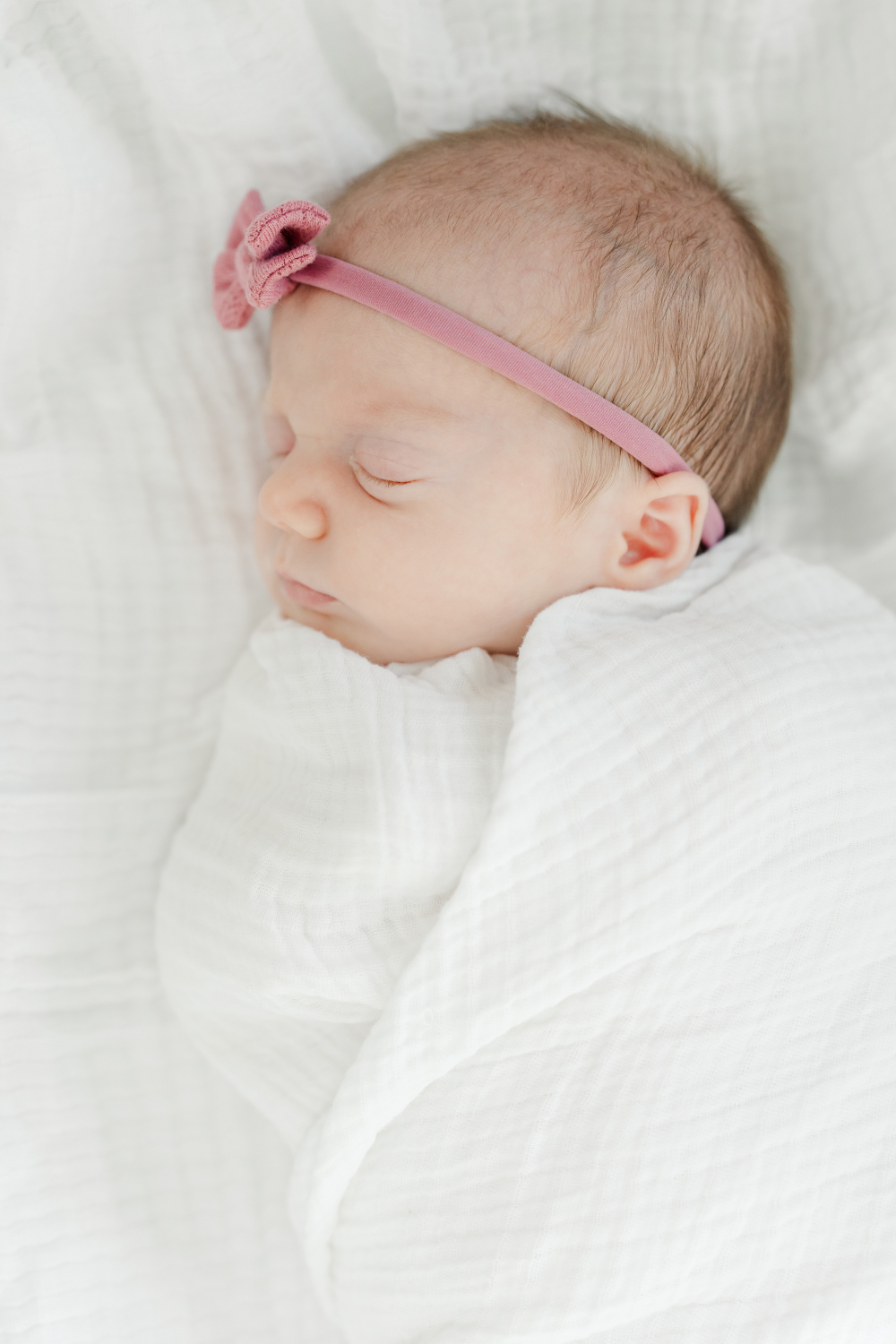 Sweet sleeping newborn wrapped in a soft white swaddle while wearing a delicate pink bow headband during an in-home session. This portrait highlights soft newborn photoshoot outfits and minimal styling that create timeless and emotional newborn photographs.