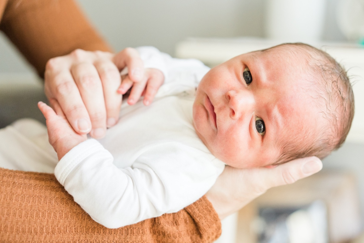 A newborn rests in a parent's arms, looking directly toward the camera with bright eyes and soft natural light. These newborn pictures highlight early personality, family connection, and the wonder of a baby's first weeks at home.