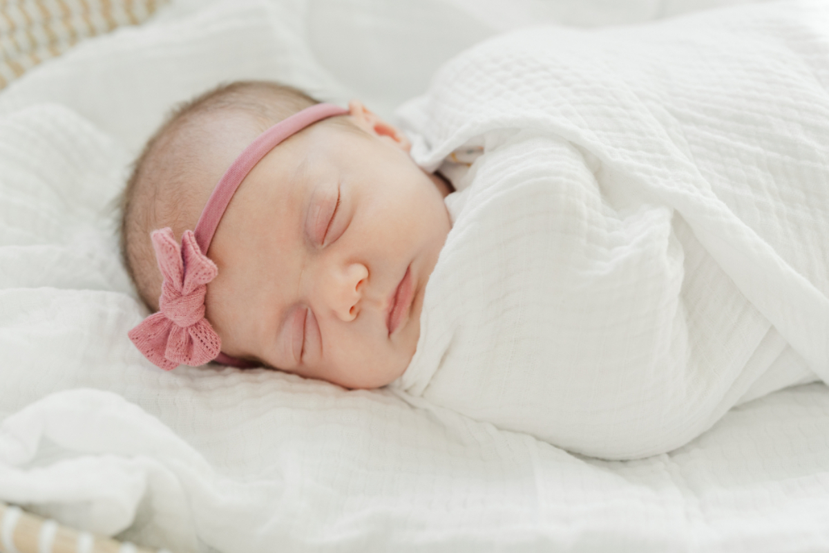 Sleeping baby girl swaddled in a soft white blanket with a pink bow headband, resting peacefully on a bed. These newborn pictures capture the sweetness, calm, and delicate details that parents never want to forget.