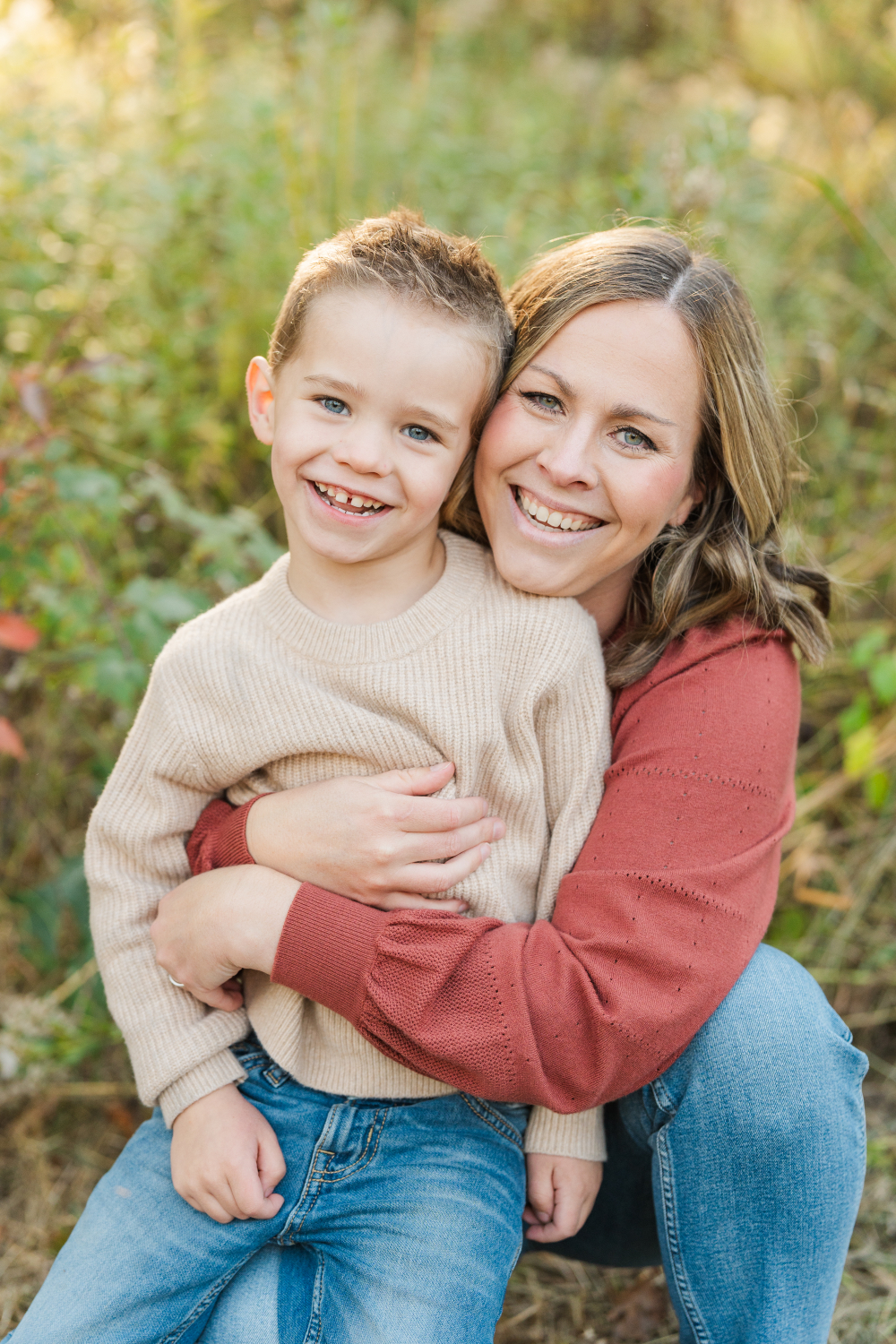 A mother hugs her young son close during an outdoor family session in Minnesota. Their genuine smiles and warm connection shine through in these golden hour photos, with soft natural light and a blurred meadow backdrop creating a timeless family portrait.