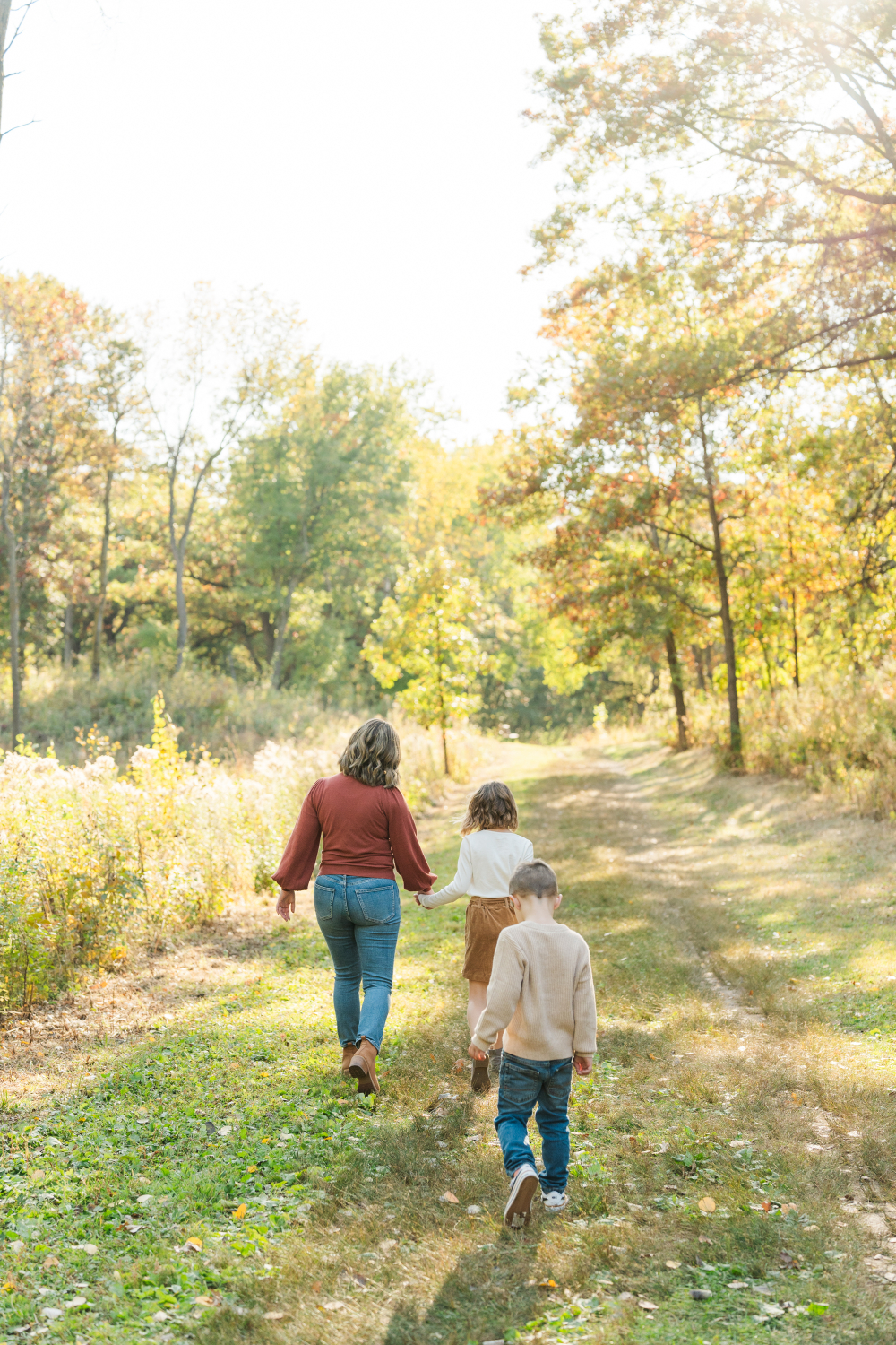 A mother walks hand in hand with her children along a peaceful tree-lined path during a family session. The soft evening light in these golden hour photos creates a dreamy atmosphere while documenting everyday moments families cherish most.