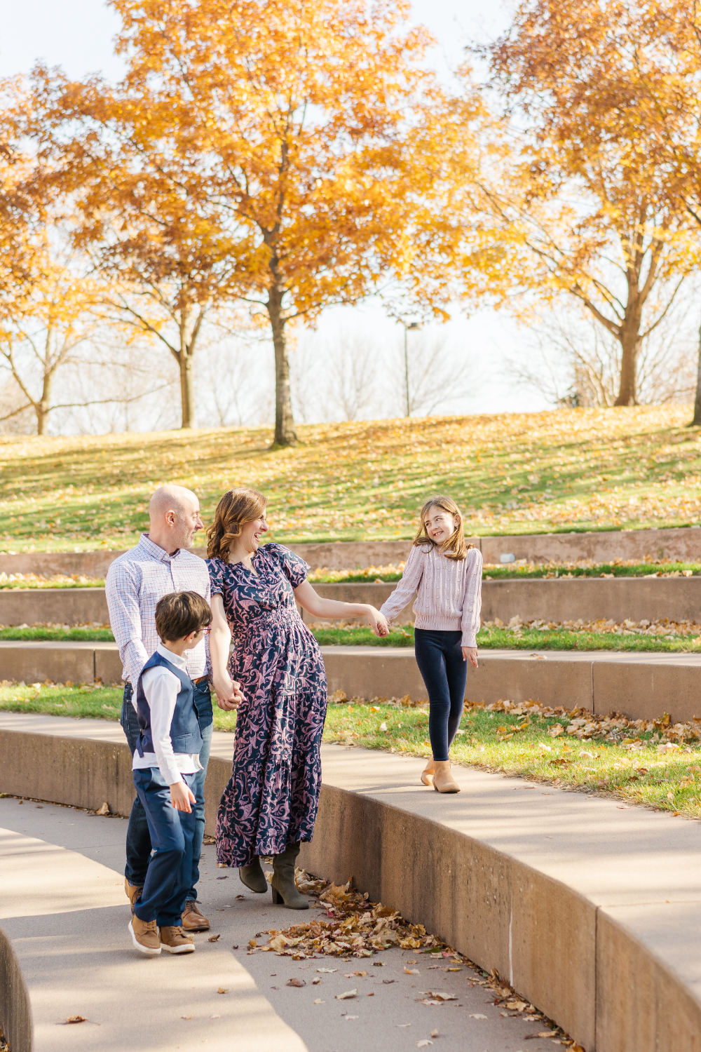 A family strolls together through a park surrounded by vibrant autumn leaves and warm sunlight. These golden hour photos highlight genuine connection and joyful interaction while showcasing the beautiful colors of a Minnesota fall evening.