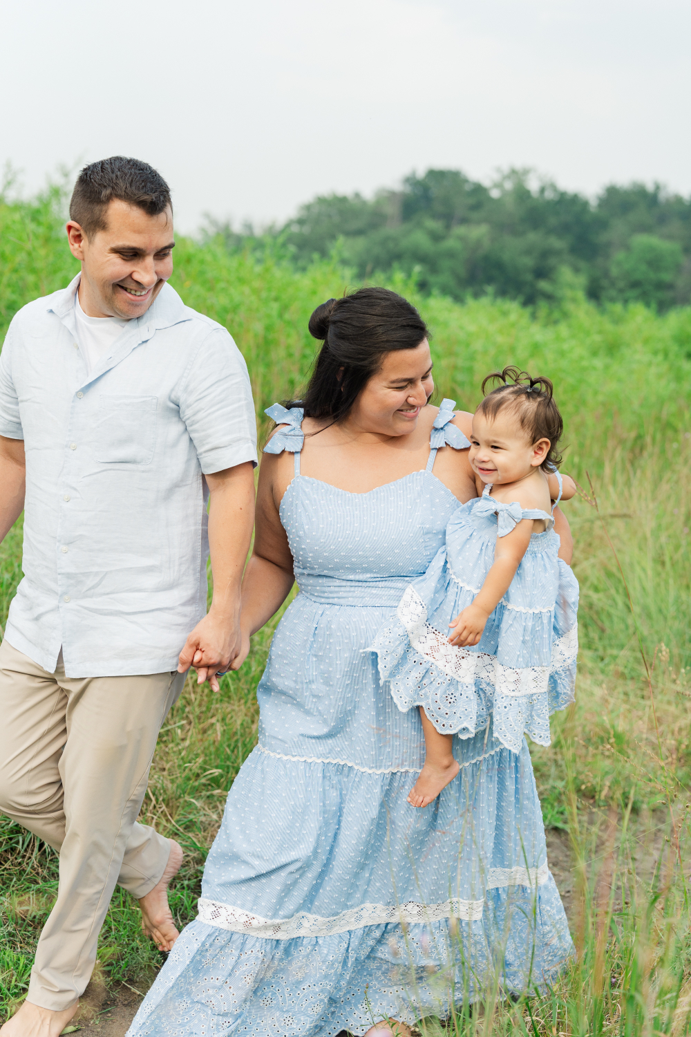 A family of three walks hand in hand through a grassy Minnesota field during an outdoor family session. Mom carries her smiling toddler while both parents share a joyful glance, capturing the connection, love, and togetherness that make family photographs so meaningful.