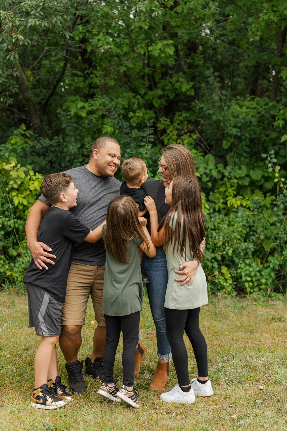 Parents and their four children gather in a group hug during a relaxed outdoor family photo session. The children laugh and lean into their parents, creating an authentic moment filled with joy, connection, and the playful energy that defines family life.