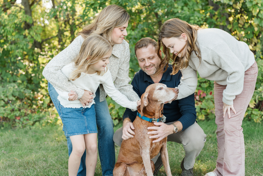 A family of four gathers around their beloved dog during a sunny outdoor portrait session. The children gently pet their furry companion while their parents smile nearby, documenting a season of life that includes every cherished member of the family.