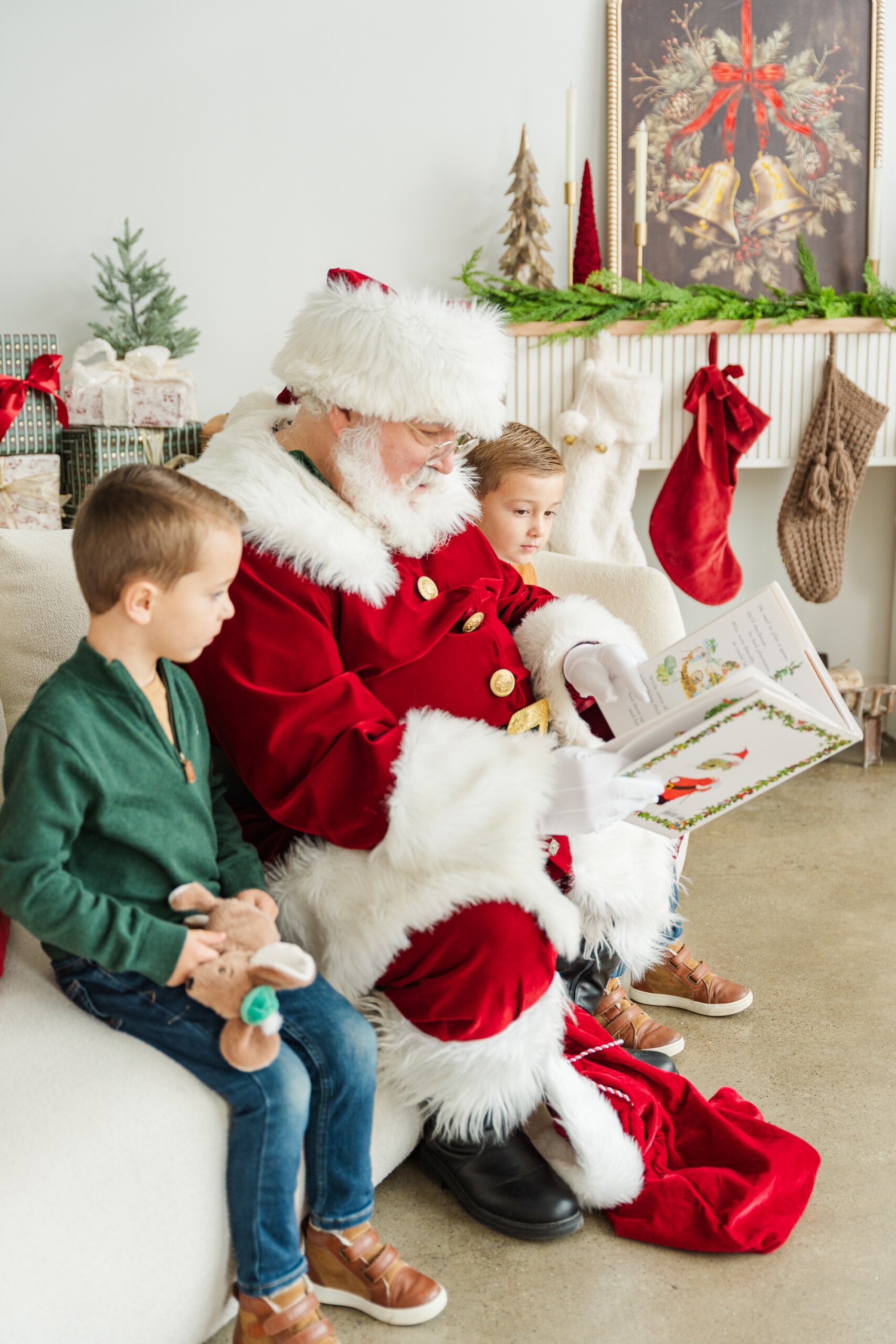 Two young boys sit beside Santa on a cozy white couch as he reads one of their favorite Christmas books for kids, surrounded by stockings, wrapped gifts, greenery, and classic red Christmas decor during a holiday photo session.